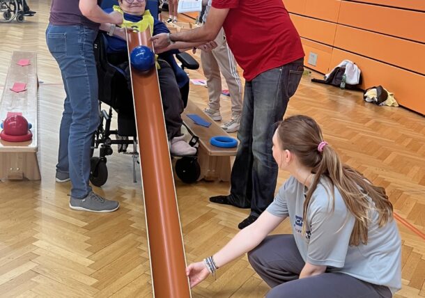Ein Kind im Rollstuhl rollt mit Hilfe von Erwachsenen in einer Turnhalle einen blauen Ball eine orangefarbene Rampe hinunter. Eine andere Frau am Ende der Rampe lenkt den Ball, und bunte Gegenstände sind als Bowlingkegel aufgestellt.