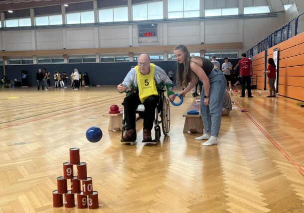 Eine junge Frau hilft einem älteren Mann im Rollstuhl, einen blauen Ball auf gestapelte rote Dosen mit den Nummern 9, 10 und 93 in einer Sporthalle zu werfen, während andere im Hintergrund zusehen und mitmachen.
