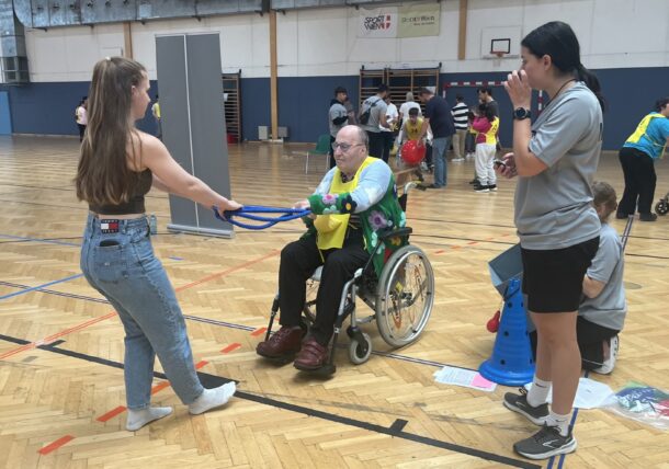 Eine junge Frau überreicht einem älteren Mann im Rollstuhl während einer Hallensportveranstaltung einen blauen Ring, während andere Teilnehmer und das Personal im Hintergrund der Sporthalle interagieren.
