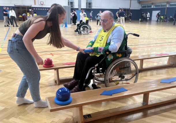 Eine junge Frau hilft einem älteren Mann im Rollstuhl während eines Spiels in einer Sporthalle. Der Mann trägt eine gelbe Weste und greift nach einem blauen Knopf auf einer Holzbank. Im Hintergrund sind weitere Personen zu sehen.