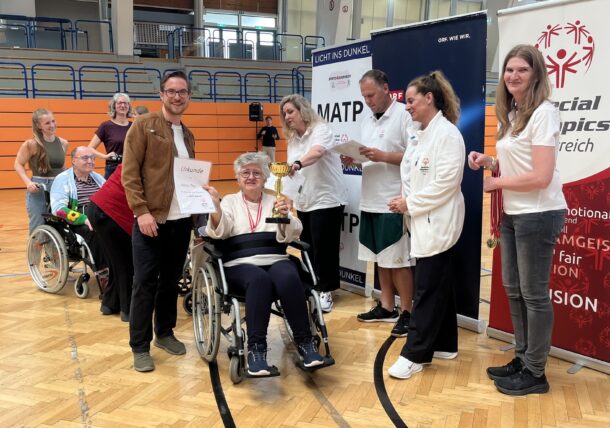Eine Gruppe von Menschen, einige davon in Rollstühlen, feiert bei einer Indoor-Veranstaltung. Ein Mann hält eine Urkunde neben einer lächelnden Frau im Rollstuhl, die eine Trophäe hält. Andere stehen in der Nähe, klatschen und lächeln. Special-Olympics-Banner sind zu sehen.