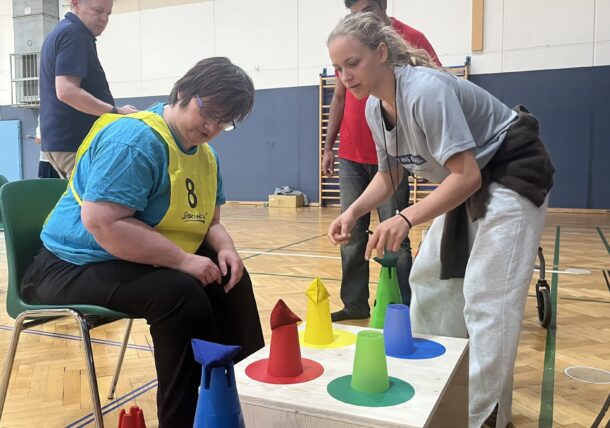 Eine Frau, die auf einem Stuhl sitzt und eine gelbe Weste trägt, nimmt an einer Fitnessaktivität teil und wird dabei von einer anderen Frau in Sportkleidung unterstützt. Sie interagieren mit bunten Kegeln auf einem weißen Kasten in einem Innenraum.