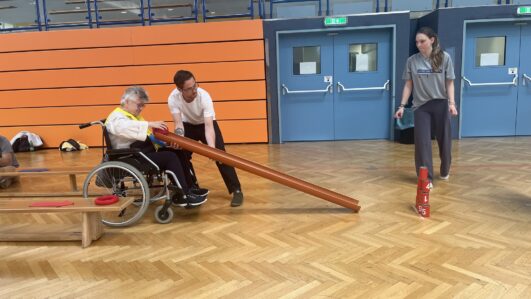 Eine ältere Person im Rollstuhl und ein Mann führen ein langes Rohr und versuchen, die in einer Turnhalle gestapelten roten Bowlingkegel umzuwerfen. Eine junge Frau schaut in der Nähe zu. Die Szene spielt auf einem hölzernen Turnhallenboden mit orangefarbenen und blauen Wänden.