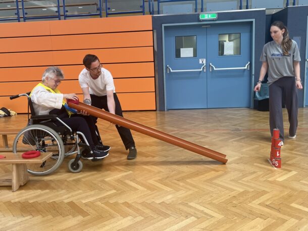 Eine ältere Person im Rollstuhl und ein Mann führen ein langes Rohr und versuchen, die in einer Turnhalle gestapelten roten Bowlingkegel umzuwerfen. Eine junge Frau schaut in der Nähe zu. Die Szene spielt auf einem hölzernen Turnhallenboden mit orangefarbenen und blauen Wänden.