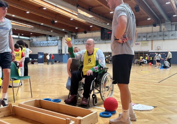 Ein älterer Mann im Rollstuhl, der eine gelbe Weste mit der Nummer 5 trägt, wirft einen kleinen gelben Ball in einer Sporthalle, umgeben von jungen Leuten, die ihm bei einer Indoor-Aktivität helfen.