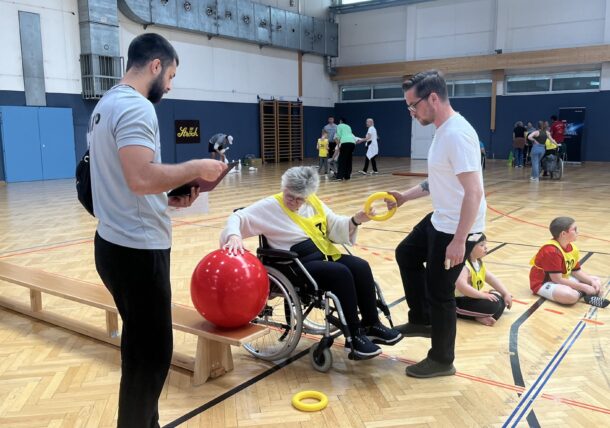 Eine Frau im Rollstuhl nimmt mit Hilfe von zwei Männern, von denen einer ihr einen gelben Ring überreicht, an einer Turnübung teil, während andere, darunter auch Kinder, im Hintergrund der Sporthalle mit verschiedenen Aktivitäten beschäftigt sind.