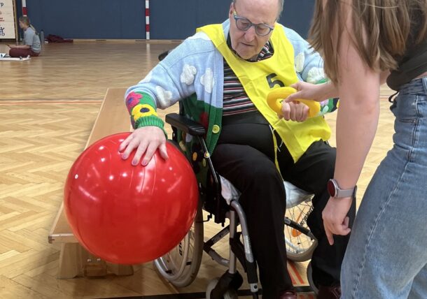 Ein älterer Mann im Rollstuhl, der eine gelbe Weste trägt, greift in einer Sporthalle nach einem großen roten Ball. Eine junge Frau steht neben ihm und bietet ihm Unterstützung und Anleitung. In der Nähe liegen gelbe Spielringe auf dem Boden.