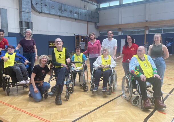 Eine Gruppe von Menschen, darunter auch Rollstuhlfahrer, die nummerierte gelbe Westen tragen, posieren und lächeln gemeinsam mit ihren Betreuern oder Unterstützern in einer Sporthalle.