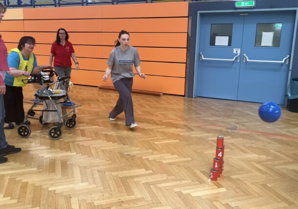 Vier Frauen spielen ein Indoor-Bowlingspiel. Eine Frau schiebt eine blaue Kugel in Richtung eines Stapels roter Becher, während die anderen zusehen und helfen. Die Szene spielt in einer Turnhalle mit Holzboden und orangefarbenen Wandpaneelen.