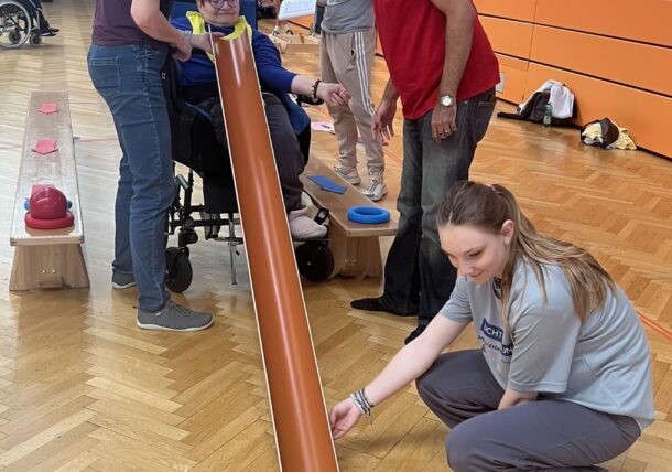 Vier Personen arbeiten in einer Turnhalle zusammen, um einen blauen Ball über eine orangefarbene Rampe zu gestapelten Dosen hinunterzurollen. Eine Person sitzt in einem Rollstuhl, zwei stehen und eine kniet, um die Rampe auszurichten. Die Atmosphäre ist kooperativ und inklusiv.