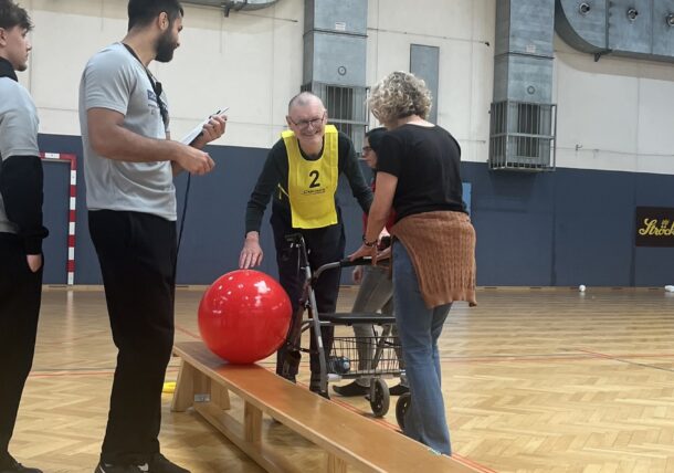Ein älterer Mann, der eine gelbe Weste trägt und eine Gehhilfe benutzt, bereitet sich darauf vor, einen großen roten Ball eine hölzerne Rampe in einer Turnhalle hinunterzurollen, wobei er von zwei Personen unterstützt und von einer weiteren Person beobachtet wird.