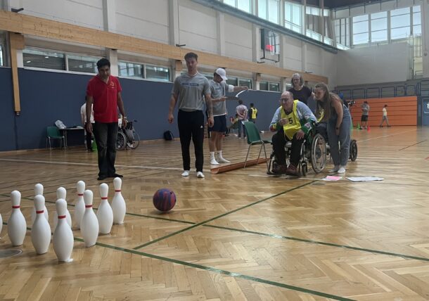 Eine Gruppe von Personen in einer Turnhalle, darunter eine Person im Rollstuhl, spielt ein adaptives Bowlingspiel mit weißen Kegeln und einer bunten Kugel auf einem Holzboden. Andere schauen zu und helfen, während die Kugel zu den Kegeln rollt.