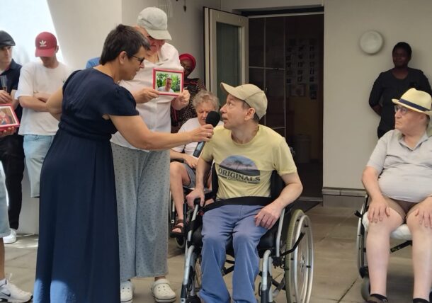 Eine Frau hält einem älteren Mann im Rollstuhl ein Mikrofon in die Hand, während andere Menschen, einige sitzend, andere stehend, zusehen und gerahmte Bilder während einer Versammlung im Freien in einer Pflegeeinrichtung halten.