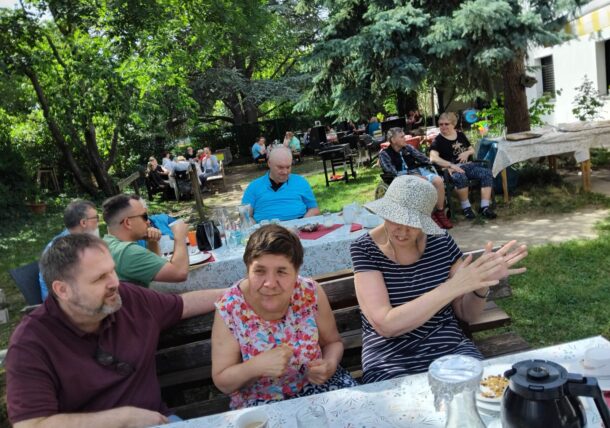 Eine Gruppe von Menschen sitzt an Tischen in einem Garten und genießt Essen und Getränke im Schatten der Bäume. Einige unterhalten sich und lächeln, und eine Frau mit Sonnenhut klatscht in die Hände. Die Atmosphäre ist locker und fröhlich.