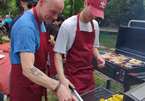 Zwei Männer mit roten Schürzen grillen Gemüse und Mais auf einem Grill bei einem Treffen im Freien. Einer benutzt eine Zange, um das Essen zu wenden, während im Hintergrund andere und Bäume zu sehen sind.