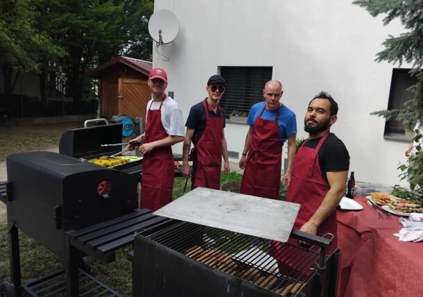 Vier Männer mit roten Schürzen stehen an Grills im Freien und kochen bei einem Treffen im Hinterhof Essen. Auf einem der Grills wird gegrillt, daneben steht ein Tisch mit Getränken und Snacks. Im Hintergrund sind ein weißes Gebäude und Bäume zu sehen.