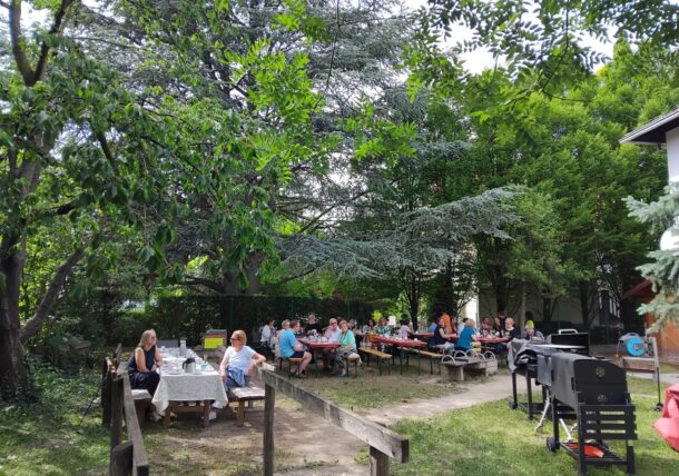 Eine Gruppe von Menschen sitzt an Picknicktischen in einem schattigen, grünen Hinterhof, der von Bäumen umgeben ist. Einige essen oder unterhalten sich, und auf einigen Tischen stehen Speisen und Getränke. Im Vordergrund ist ein Grill zu sehen.