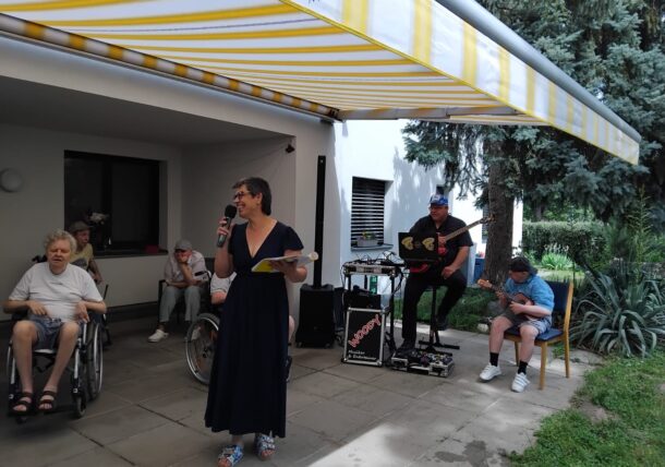 Eine Frau, die ein Mikrofon und Papiere in der Hand hält, spricht im Freien zu einer Gruppe, zu der auch Rollstuhlfahrer gehören, in der Nähe einer Band mit zwei Musikern, die unter einer gelb gestreiften Markise neben einem Gebäude und einer Grünanlage Gitarre spielen.