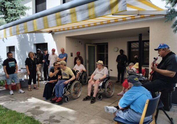 Eine Gruppe von Menschen, einige in Rollstühlen, andere stehend, versammelt sich im Freien unter einer gelb gestreiften Markise. Eine Person spielt Gitarre, während andere zuschauen und sich an einem sonnigen Tag in der Nähe eines Gebäudeeingangs treffen.