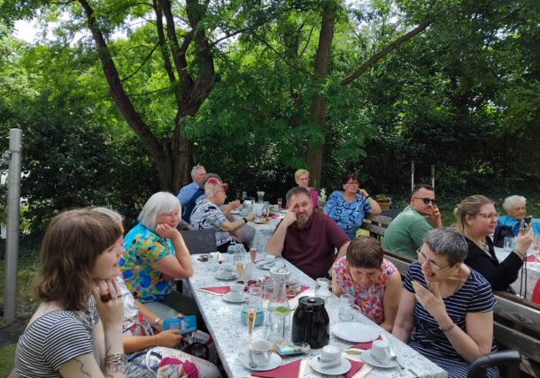 Eine Gruppe von Menschen sitzt an Tischen im Freien unter Bäumen und teilt eine Mahlzeit mit Kaffeetassen, Tellern und Getränken. Die Umgebung wirkt entspannt und gesellig, mit viel Grün und Schatten von den umliegenden Bäumen.