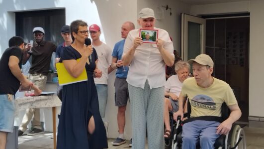 Eine Gruppe von Menschen, darunter eine Frau, die in ein Mikrofon spricht, ein Mann im Rollstuhl und ein weiterer Mann, der ein gerahmtes Foto in der Hand hält, versammelt sich an einem sonnigen Tag im Freien vor einem weißen Gebäude.