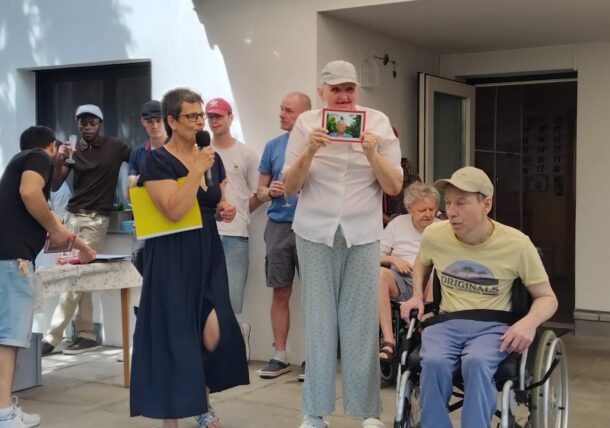 Eine Gruppe von Menschen, darunter eine Frau, die in ein Mikrofon spricht, ein Mann im Rollstuhl und ein weiterer Mann, der ein gerahmtes Foto in der Hand hält, versammelt sich an einem sonnigen Tag im Freien vor einem weißen Gebäude.