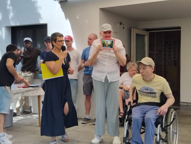 Eine Gruppe von Menschen, darunter eine Frau, die in ein Mikrofon spricht, ein Mann im Rollstuhl und ein weiterer Mann, der ein gerahmtes Foto in der Hand hält, versammelt sich an einem sonnigen Tag im Freien vor einem weißen Gebäude.