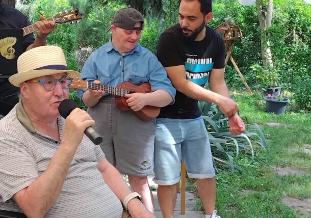 Ein älterer Mann im Rollstuhl singt im Freien in ein Mikrofon, während ein jüngerer Mann und ein Mensch mit Down-Syndrom Instrumente spielen und lächeln. Bäume und Lichterketten schmücken den Hintergrund.