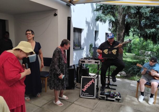 Eine Gruppe von Menschen genießt Live-Musik im Freien. Ein Mann spielt Gitarre neben einer Musikanlage mit der Aufschrift Woopy, während andere singen und lächeln. Eine Frau in einem roten Kleid hält ein Mikrofon, und eine andere Person spielt ein kleines Instrument.