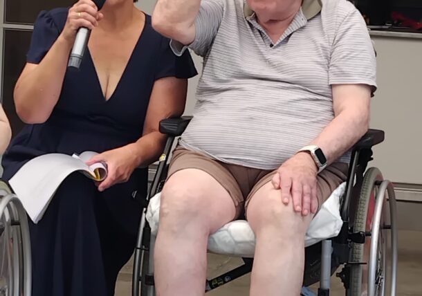 Eine Frau in einem dunklen Kleid, die ein Mikrofon hält, sitzt neben einem älteren Mann im Rollstuhl, der einen Hut und eine Brille trägt, seinen Hut berührt und lächelt. Sie scheinen bei einer Veranstaltung in einem Gebäude oder im Freien zu sein.
