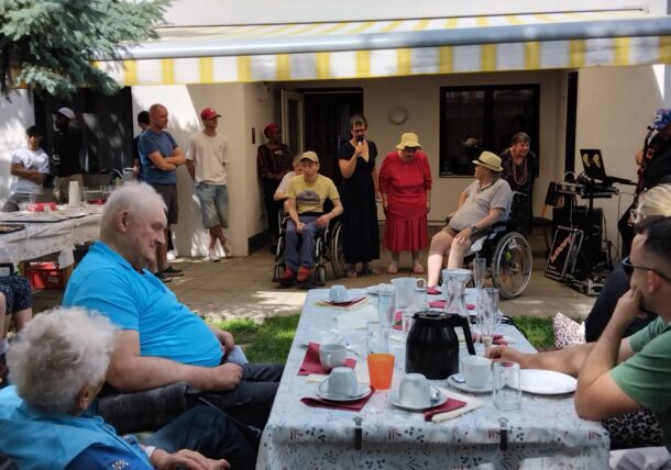 Eine Gruppe von Menschen, darunter mehrere Rollstuhlfahrer, versammelt sich im Freien um einen gedeckten Tisch, um Tee zu trinken. Einige stehen, andere sitzen unter einer gelb gestreiften Markise und hören einem Sprecher zu. Im Hintergrund sind Musikanten und Schaulustige zu sehen.