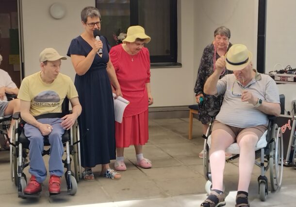 Vier ältere Menschen, drei Frauen und ein Mann, unterhalten sich an einem sonnigen Tag im Freien mit einem Mann mit Hut, der in einem Rollstuhl sitzt. Eine Frau hält ein Mikrofon, die anderen schauen zu und lächeln.