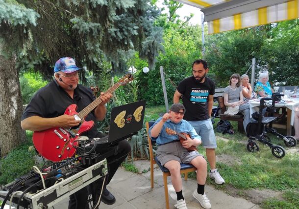 Ein Mann spielt eine E-Gitarre und singt im Freien unter einem Vordach. In der Nähe stehen ein sitzendes Kind und ein Erwachsener zusammen, während mehrere Personen an einem Tisch im Hintergrund sitzen und das Beisammensein in einem grünen Garten genießen.