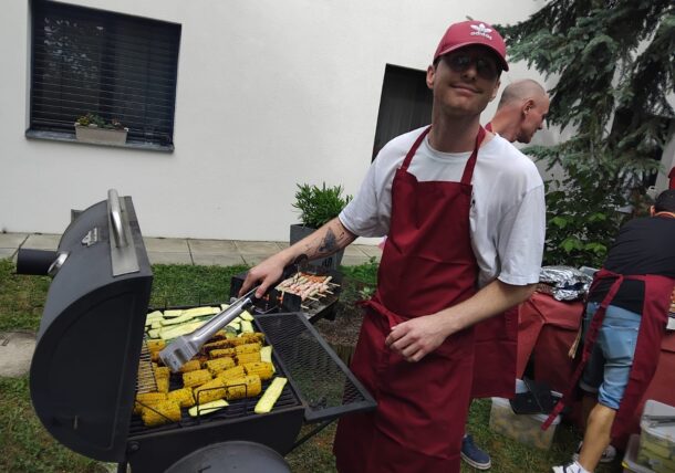 Ein Mann in roter Schürze und Mütze grillt Mais und Zucchini im Freien und lächelt in die Kamera. Eine andere Person in roter Schürze bereitet an einem Tisch in der Nähe das Essen zu. Schauplatz ist ein Hinterhof mit Grünpflanzen und einem weißen Gebäude.