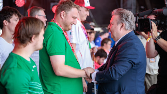 Ein Mann im Anzug schüttelt auf der Bühne einem Mann in einem grünen Hemd die Hand, umgeben von anderen Menschen. Ein Kameramann filmt die Szene. Rote Bühnenlichter sorgen für eine festliche Atmosphäre.