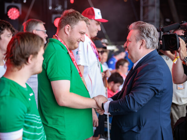 Ein Mann im Anzug schüttelt auf der Bühne einem Mann in einem grünen Hemd die Hand, umgeben von anderen Menschen. Ein Kameramann filmt die Szene. Rote Bühnenlichter sorgen für eine festliche Atmosphäre.