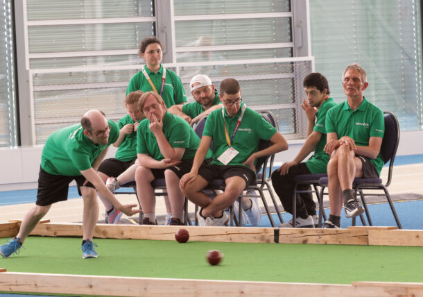 Eine Gruppe von Menschen in grünen T-Shirts schaut zu, wie eine Person auf einem Boccia-Platz in der Halle einen Ball rollt. Einige sitzen, andere stehen, doch alle verfolgen das Spiel mit großer Aufmerksamkeit.
