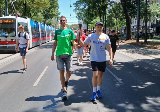 Zwei Männer gehen Seite an Seite eine sonnige Straße entlang, halten eine Fackel in der Hand und führen eine Gruppe von Menschen an. Im Hintergrund säumen eine rote Straßenbahn und Bäume die Straße.