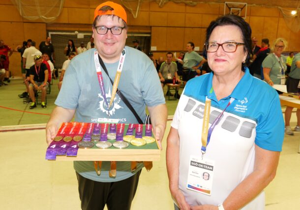 Ein lächelnder Mann in einem blauen Special-Olympics-T-Shirt und einer orangefarbenen Mütze hält ein Tablett mit Medaillen in der Hand. Neben ihm steht eine Frau in einem blau-weißen T-Shirt. Beide befinden sich in einer Halle bei einer Sportveranstaltung, im Hintergrund sind Menschen zu sehen.
