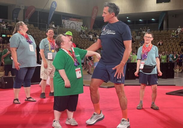 Ein Mann in Sportkleidung lächelt und hält auf einer roten Matte die Hand eines Special-Olympics-Athleten, umgeben von anderen fröhlichen Teilnehmern in einer Sporthalle.