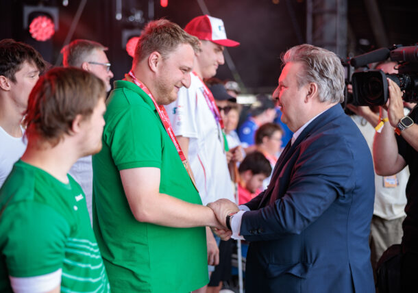 Ein Mann im Anzug schüttelt auf der Bühne einem Mann in einem grünen Hemd herzlich die Hand, umgeben von anderen Menschen und einem Kameramann, der diesen Moment festhält.