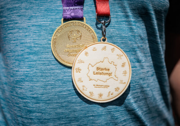Um den Hals der Person hängen zwei Medaillen: eine goldene mit der Aufschrift „Special Olympics“ und eine aus Holz mit der Gravur „Starke Leistung!“, umgeben von kleinen Sport-Symbolen. Die Person trägt ein blaues T-Shirt.