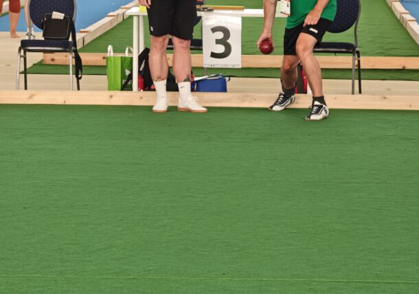 Zwei Männer in grünen Hemden spielen drinnen Boccia. Der eine ist gerade dabei, eine rote Kugel zu werfen, während der andere zusieht. Drei rote und zwei grüne Kugeln liegen verstreut neben einer kleinen weißen Zielkugel auf der grünen Matte.