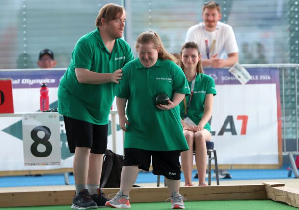 Zwei Sportler in grünen Trikots lächeln, während sie bei einer Sportveranstaltung Boccia spielen. Der eine hält eine Boccia-Kugel in der Hand, der andere feuert ihn sanft an. Im Hintergrund schauen Zuschauer zu.