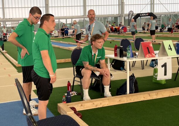 Vier Männer in grünen Trikots, vermutlich Mannschaftskameraden, verfolgen bei einer Hallensportveranstaltung gebannt ein Spiel. Ein Mann sitzt, während die anderen daneben stehen. Auf dem grünen Spielfeld sind Anzeigetafeln und Boccia-Kugeln zu sehen.