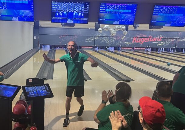 Ein Mann in einem grünen Hemd feiert auf einer Bowlingbahn, während Menschen in passenden Hemden klatschen und jubeln. Im Hintergrund sind Bowlingkugeln und elektronische Anzeigetafeln zu sehen.