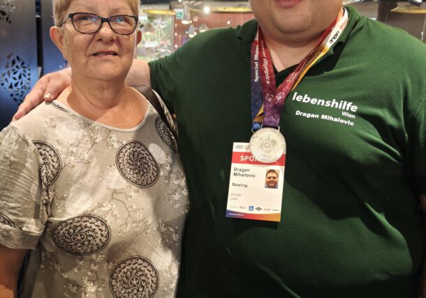 Ein lächelnder Mann, der Orden und ein Namensschild trägt, steht neben einer älteren Frau mit Brille. Sie befinden sich in einem Raum, stehen eng beieinander und blicken beide in die Kamera.