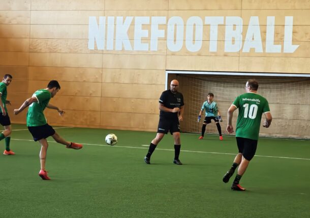 Vier Männer in grünen Fußballtrikots spielen in der Halle auf Kunstrasen, während ein Mann in Schwarz als Schiedsrichter fungiert und ein weiterer Mann in Schwarz als Torwart steht. An der Holzwand hinter dem Tor befindet sich ein großes NIKEFOOTBALL-Schild.