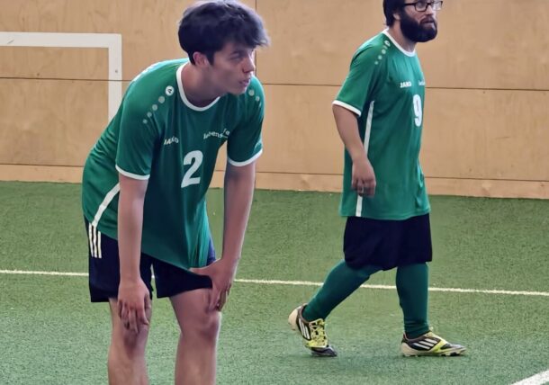 Zwei Männer in grünen Fußballtrikots stehen auf einem Kunstrasenplatz in der Halle. Der Mann links mit der Rückennummer 2 beugt sich mit den Händen auf den Knien nach vorne, während der Mann rechts mit der Rückennummer 9 aufrecht steht.