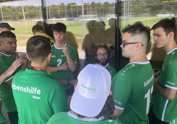 Eine Gruppe von Männern in grünen „Lebenshilfe Wien“-Trikots steht in einer kleinen Runde beieinander, unterhält sich und hört aufmerksam zu – in der Nähe eines Fensters, durch das man einen Sportplatz sehen kann.